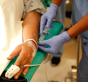 Nahaufnahme einer Krankenpflegekraft, die ein Medikament in eine Kanüle an der Hand eines Mannes injiziert.